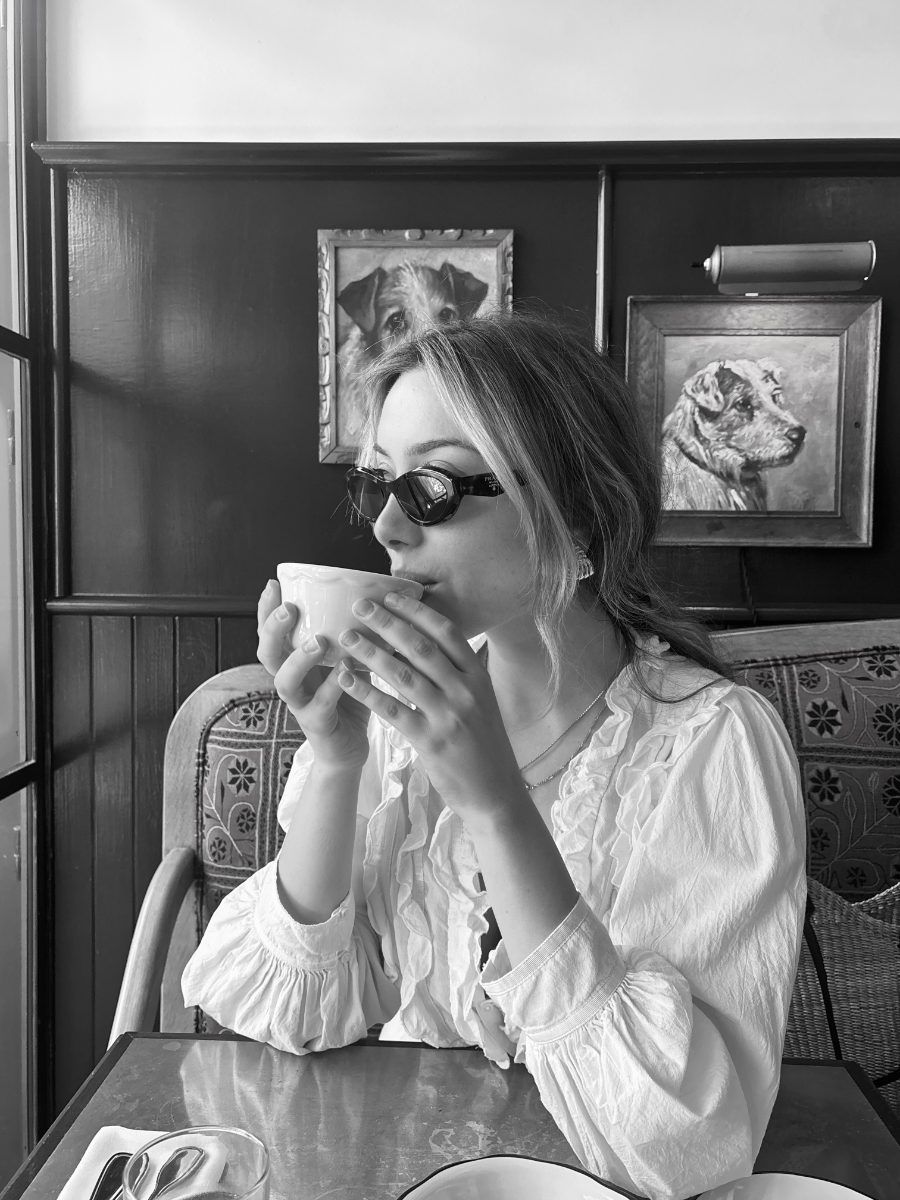 Black and white photograph of Andrea Manokian sitting in a diner booth, drinking from a cup, framed pictures on the wall behind her.