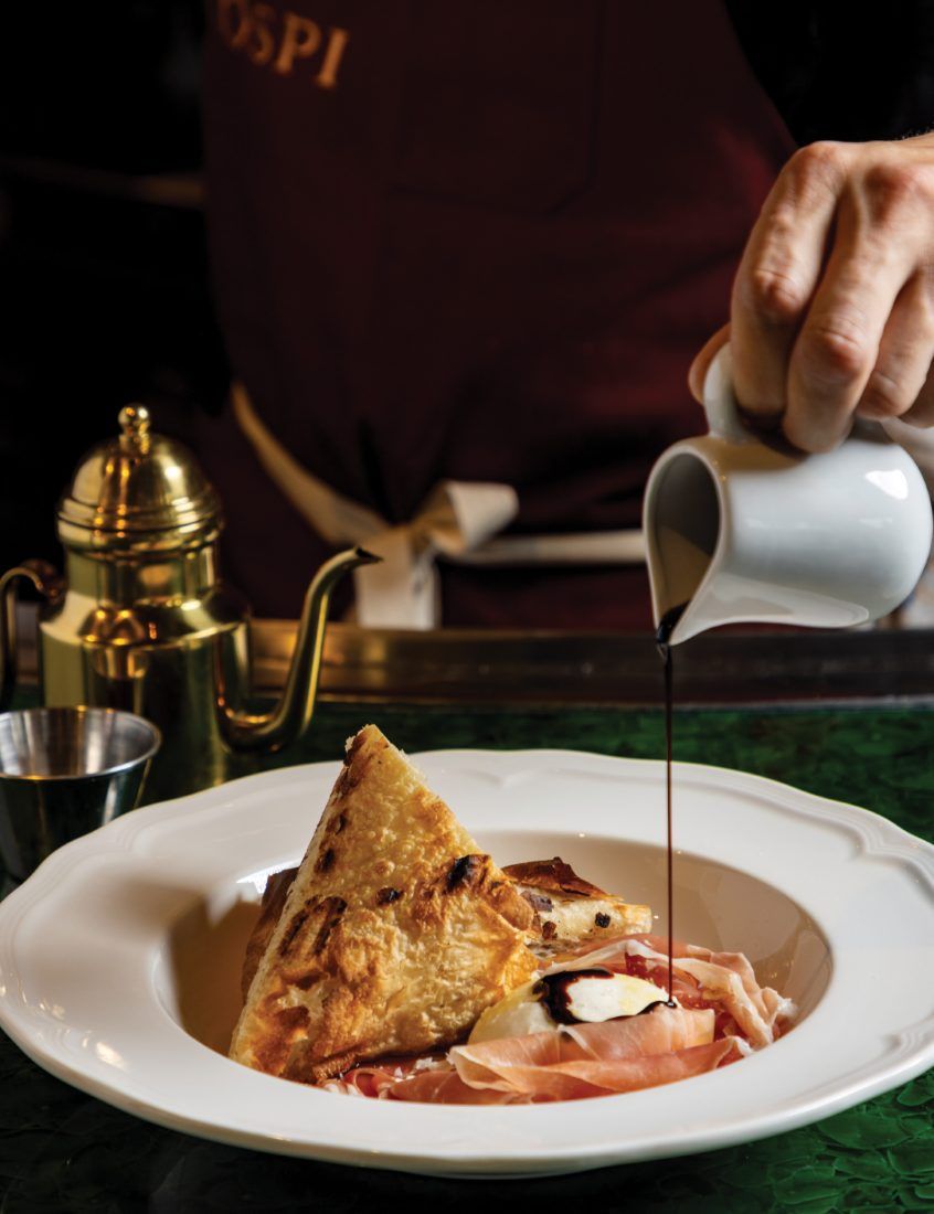 A hand pouring sauce from a small jug over a plated dish, beside a brass teapot on a dark table.