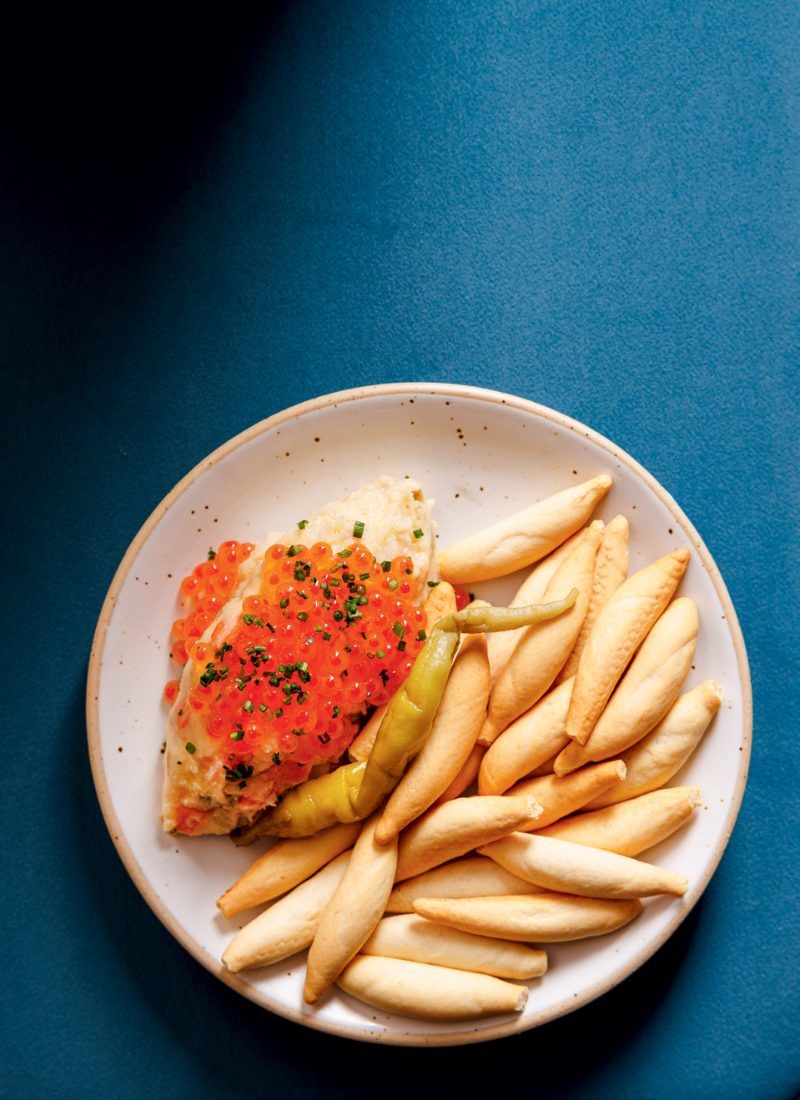 A white plate of pale fried potatoes topped with salmon roe, chives and a green chilli, on a deep blue surface.