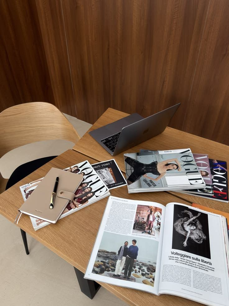 Wooden desk with an open laptop, a notebook and pen, and magazine spreads fanned out across the surface.