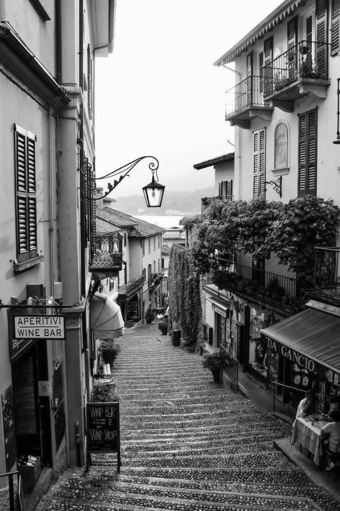 Black and white photograph of a narrow stepped European street lined with shuttered buildings, a wine bar sign and hanging lamps.